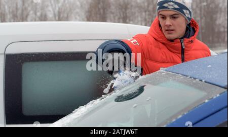 Ein junger Mann räumt Schnee aus einem Auto, ein junger Mann räumt am 7. Januar 2019 das Autoglas aus Schnee, Gebiet von Moskau, Russland Stockfoto