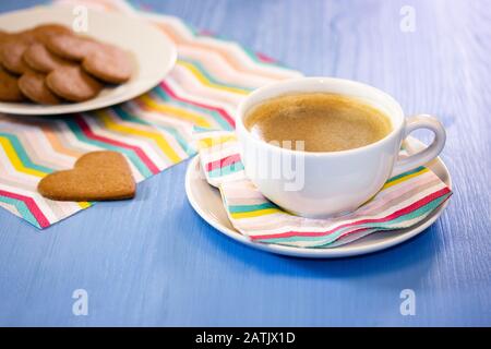Weiße Tasse Kaffee mit Plätzchen auf einem blauen Tisch mit gestreiften Servietten. Morgens schwarzer heißer Kaffee mit hausgemachtem süßem Gebäck und herzförmigem Lebkuchen Stockfoto