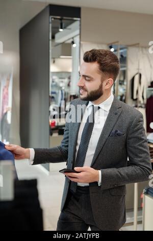 Lächelnder, stilvoller junger Mann in der Jacke, der im Rack steht und das Smartphone hält, während er Kleidung im Geschäft kauft Stockfoto