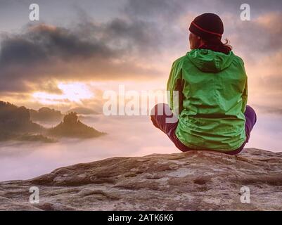 Grüne Jacke Frau sitzt penibel am Rand eines Felsens und betrachtet die nebligen Wolken. Stockfoto