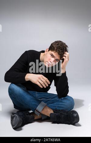 Stilvoller Kerl, der im Studio sitzt und in Denimhosen und schwarzem Sweatshirt sitzt. Männliches Studentenporträt voll auf weißem Hintergrund. Studio gedreht Stockfoto