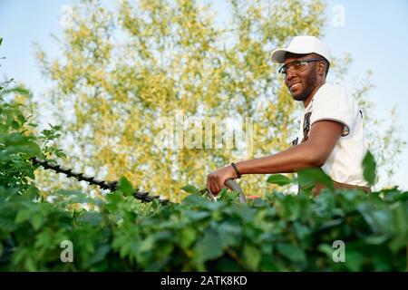 Afrikanischer Gärtner mit Overalls und Schutzbrille, die mit professioneller Gartenausrüstung arbeiten. Fröhlicher junger Mann, der Büsche fräst. Saisonarbeitskonzept Stockfoto