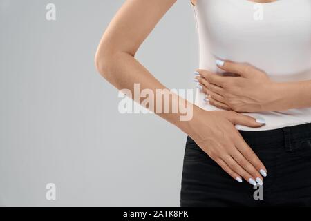 Clolse von Incognito kaukasische Frau in lässiger Anzug posiert über Grau isoliert Hintergrund und berühren Magen wegen der Leber Ache. Crop von brunette leiden unter Bauchschmerzen. Stockfoto