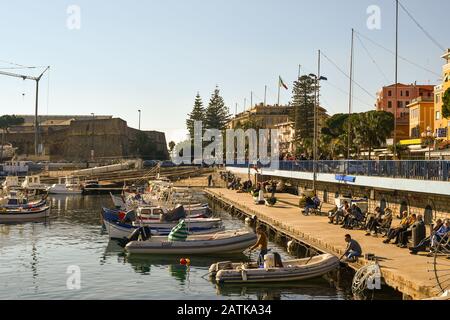Blick auf den Alten Hafen mit Menschen und Touristen, die auf der Promenade spazieren gehen und die Sonne genießen, die auf den Bänken des Kais sitzt, Sanremo, Ligurien Italien Stockfoto