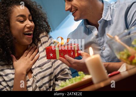 Romantisches Datum. Junges multiethnisches Paar zu Hause sitzend am Tisch essen Ehemann Abendessen, das Geschenk an Frau, die die Brust berührt, überraschte Nahaufnahme Stockfoto