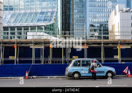 Eine Taxifahrerin steigt in ihr Taxi, das vor Bauarbeiten geparkt ist, und ist auf modernen Büroblöcken ausfahrend. Stockfoto