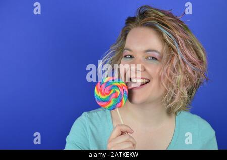 Porträt einer jungen blonden Frau mit bunten Haaren, die sich an einem großen Lollipop leckt und in die Kamera schaut Stockfoto