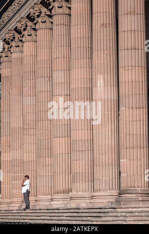 Ein Mann steht neben den massiven Steinsäulen aus rosafarbenem Steinbruch des staatlichen Regierungspalasts und Museums oder des Palacio de Gobierno del Estado de Nuevo Leon im Macroplaza Grand Plaza neben dem Barrio Antiguo Viertel Monterrey, Nuevo Leon, Mexiko. Stockfoto