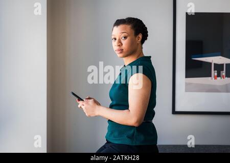 Seitenansicht der Geschäftsfrau Textnachrichten auf dem Smartphone, während sie im Büro an der Wand steht Stockfoto