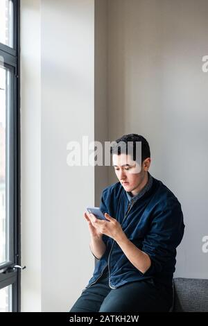 Junger Geschäftsmann, der mit einem Smartphone auf dem Sofa gegen die Wand im Büro sitzt Stockfoto