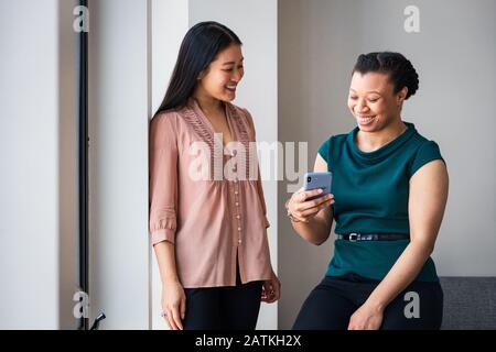 Fröhliche weibliche Kollegen, die das Smartphone gegen die Wand im Büro betrachten Stockfoto