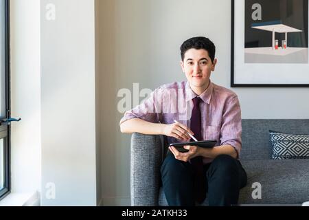Porträt eines selbstbewussten Geschäftsmannes mit Grafiktablett, das auf dem Sofa gegen die Wand im Büro sitzt Stockfoto