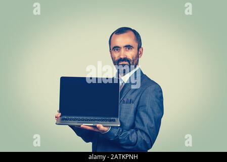 Bärtiger Geschäftsmann mit leerem schwarzem Notebook-Bildschirm. Mixed Race Bärtiermodell isoliert auf grün-gelbem Studiohintergrund mit Kopierraum. Horizont Stockfoto