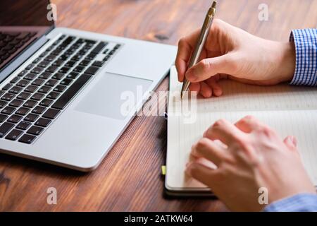 Ein Mann notiert sich in einem Tagebuch neben einem Laptop auf einem Holztisch. Nahaufnahme. Stockfoto