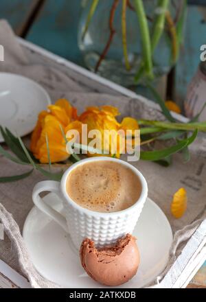 Nahansicht einer Tasse Kaffee mit einem Stück Makkaron auf der Kaffeeschale. Ein paar orangefarbene Butterbecher befinden sich auf der Rückseite der Szene. Stockfoto