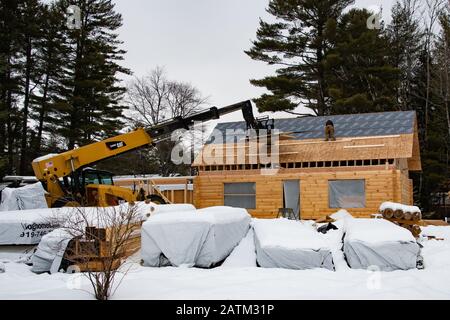 Zwei Bauarbeiter, die im Winter eine Dachabdeckung auf einem Holzhaus mit einem Cat TL1055C Teleskoplader in Speculator, NY USA, installieren. Stockfoto