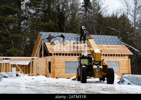 Zwei Bauarbeiter, die im Winter eine Dachabdeckung auf einem Holzhaus mit einem Cat TL1055C Teleskoplader in Speculator, NY USA, installieren. Stockfoto