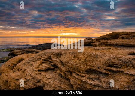 Unter den Strahlen der aufgehenden Sonne sind orangefarbene Sandstrandfelsen eingefärbt. Stockfoto