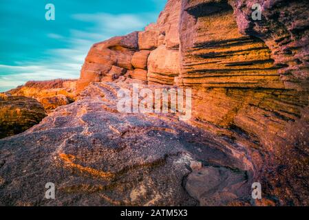 Unter den Strahlen der aufgehenden Sonne sind orangefarbene Sandstrandfelsen eingefärbt. Stockfoto