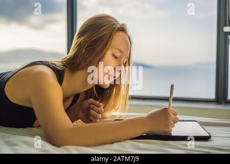 Junge Frau zieht vor dem Hintergrund eines Fensters mit Meerblick auf eine Tablette. Designer, Skizzen Stockfoto