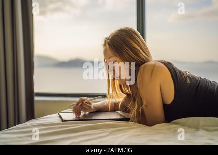 Junge Frau zieht vor dem Hintergrund eines Fensters mit Meerblick auf eine Tablette. Designer, Skizzen Stockfoto