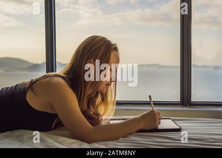 Junge Frau zieht vor dem Hintergrund eines Fensters mit Meerblick auf eine Tablette. Designer, Skizzen Stockfoto