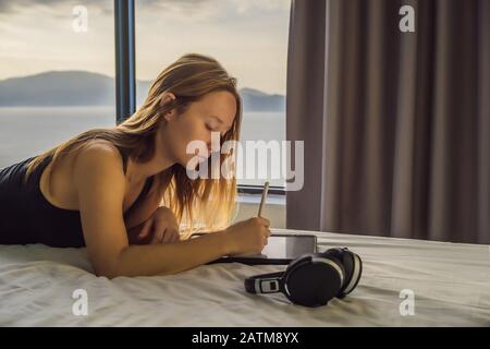 Junge Frau zieht vor dem Hintergrund eines Fensters mit Meerblick auf eine Tablette. Designer, Skizzen Stockfoto