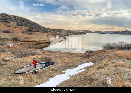 Senioren-Paddler in einem trockenen Anzug und einer Schwimmweste trägt sein Paddleboard an einem Ufer des Horsetooth Reservoirs im Norden Colorados, Winterpaddeln Stockfoto