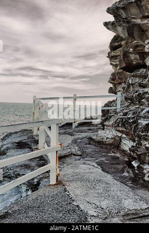 Bronte Beach mit Blick auf den Südpazifik am Ende des Weges, vorbei am Pool, schroffen Klippen, weißen Holzpfosten und Schienenzaun Stockfoto