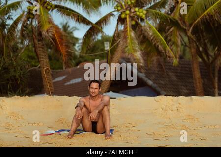 Ein junger Erwachsener sitzt auf dem Sand des Karon Beach in Thailand auf einem Grund von Kokospalmen und einem Hausdach Stockfoto