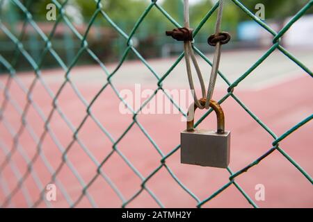 Nahaufnahme von verrostetem Vorhängeschloss am grünen Zaun mit Kabelanschluss. Tennisplatz im Hintergrund, Stockfoto