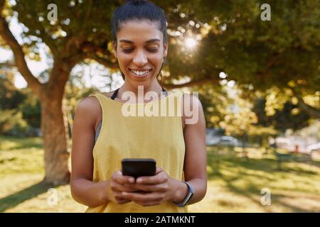 Portrait einer lächelnden jungen, sportlichen Frau, die im Park mit dem Handy telefoniert, bevor sie Sport macht Stockfoto