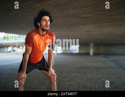 Portrait eines müden kaukasischen Sportlers, der Musik hört, die nach einem langen Lauf unter der Brücke atmen kann Stockfoto