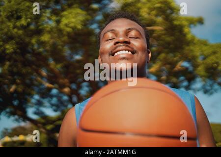 Porträt eines lächelnden, zufriedenen jungen männlichen Spielers mit geschlossenen Augen, der Basketball im Freien hält Stockfoto