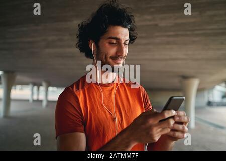 Porträt eines lächelnden jungen, sportlichen Mannes, der mit dem Schreiben auf einem Smartphone beschäftigt ist, das unter der Brücke steht Stockfoto