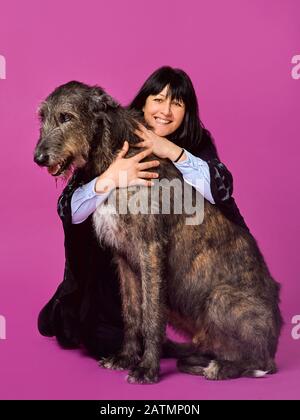 Lächelnde fröhliche brünette Frau mit grauen irischen Wolfhounds auf fuchsienfarbenem Hintergrund im Fotostudio. Freundschaft, Liebe, Haustierkonzept. Stockfoto