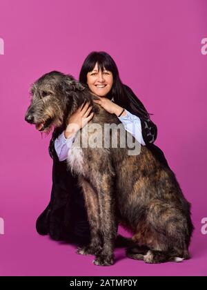 Lächelnde fröhliche brünette Frau mit grauen irischen Wolfhounds auf fuchsienfarbenem Hintergrund im Fotostudio. Freundschaft, Liebe, Haustierkonzept. Stockfoto