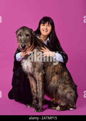 Lächelnde fröhliche brünette Frau mit grauen irischen Wolfhounds auf fuchsienfarbenem Hintergrund im Fotostudio. Freundschaft, Liebe, Haustierkonzept. Stockfoto