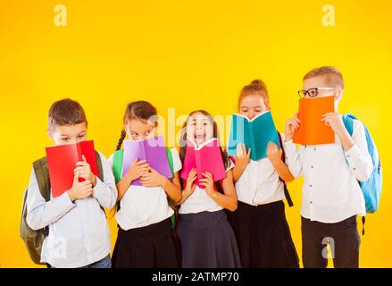 Fröhliche Schulkinder stehen mit bunten Büchern isoliert auf gelbem Hintergrund. Kinder verstecken sich hinter Büchern Stockfoto