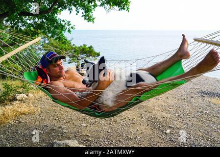 Ein junger Mann ruht mit einem Hund in einer Hängematte am Strand Stockfoto