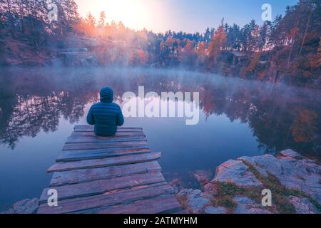 Ein Mann, der auf einem Holzdeck sitzt und einen See mit einem Ufer aus Granit betrachtet. Sonnenaufgang über dem See Stockfoto