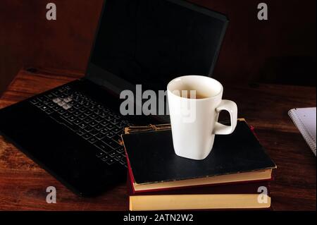 Horizontales Foto von weißem Kaffeebecher auf einem Stapel alter Bücher und Alben auf einem alten, zerkratzten dunkelbraunen Tisch. Laptop im Hintergrund. Stockfoto
