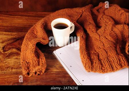 Brauner Stricktedd-Pullover mit Flechten, die einen weißen Becher mit Kaffee umfassen. Notizbuch liegt unter dem Kleidungsstück. Stockfoto