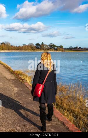 Eine blonde Frau in einem schwarzen Mantel mit rotem Beutel läuft auf dem Solent Way Fußweg bei Langstone, in der niedrigen Wintersonne bei Flut, Hampshire, Großbritannien Stockfoto