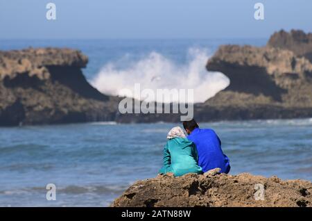 Ein junges muslimisches Liebespaar sitzt auf den Felsen am Meeresrand und beobachtet, wie die hohen Wellen gegen weit entfernte Felsen abstürzen. Marokko, Nordafrika. 2020. Stockfoto