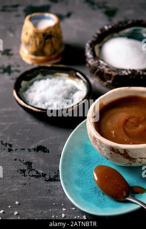 Hausgemachte gesalzene Karamellsoße mit Fleur de sel-Salz. Zutaten in Keramikschalen oben. Grauer strukturierter Leinwandhintergrund. Stockfoto