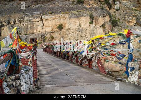 Stahlbrücke mit Holzboden mit buddhistischen Gebetflaggen, die im Wind über den Fluss im Himalaya in der Nähe von Kada, Himachal Pradesh, Indien, flattern. Stockfoto