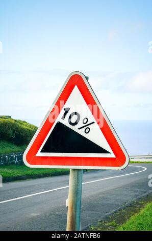 Rotes Dreieck mit Straßenschild, das auf eine steile Steigung von 10 Prozent auf der Straße hinweist. Leere Straße umgeben von grünem Gras im Hintergrund. Verkehrszeichen. Vertikales Foto. Stockfoto