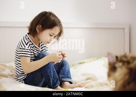 Süßes kleines Mädchen, das allein auf einem Bett sitzt und mit einer Katze spielt, die mit ernstem Gesicht, seitlichem Blick und voller Länge spielt Stockfoto
