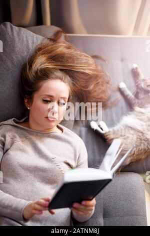 Attraktives Weibchen mit langem Haar Lesen interessantes Buch, mit Katze zu Hause ruhen, Draufsicht Stockfoto